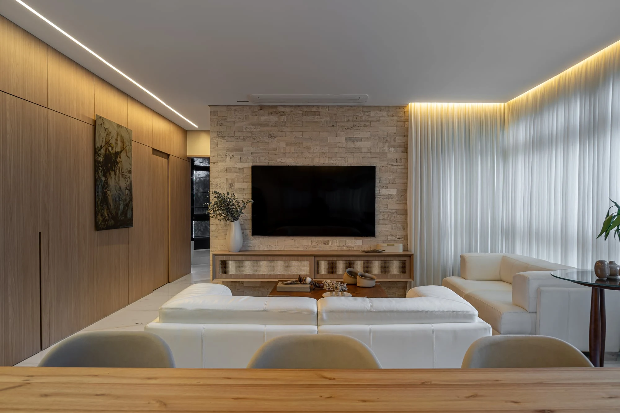 Sala de estar e jantar integradas, com sofá branco, painel de madeira e cortinas leves